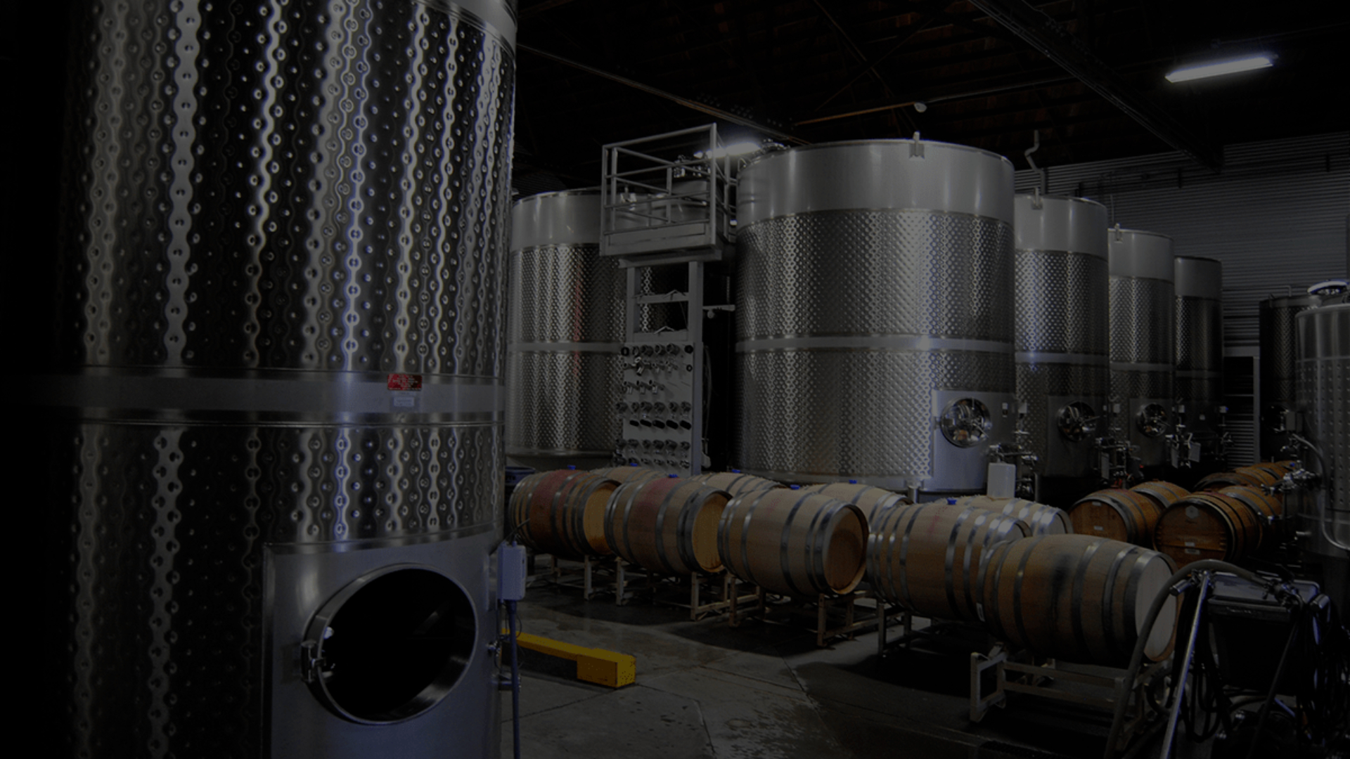 A dark room filled with winery fermentation tanks with wooden wine barrels on stands in between the tanks.