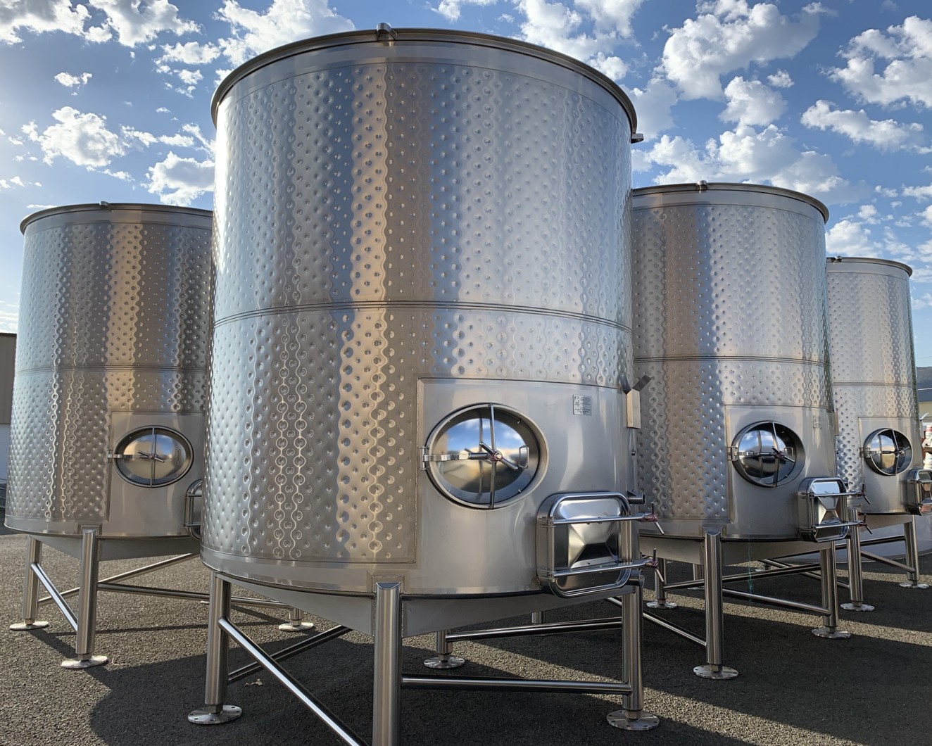 Four large medium sized winery fermentation tanks outside with blue sky and white fluffy clouds.
